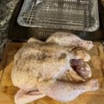 Raw whole chicken seasoned with spices on a wooden cutting board with a cooling rack in background.
