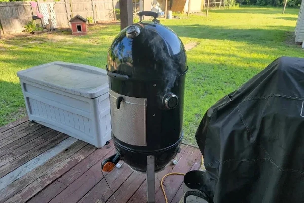 Black rounded outdoor smoker or grill with silver panel, gauges, wheels, and top vent on a wooden deck.