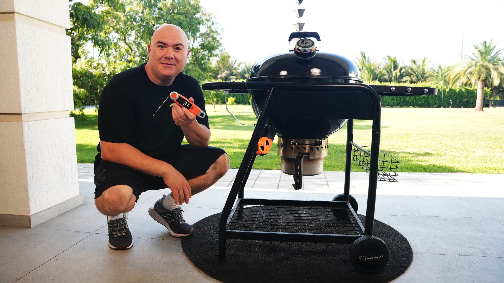 Black kettle-style grill with domed lid and temperature gauge stands on a patio beside a lawn.
